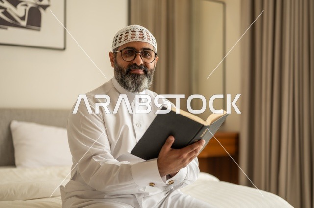 Spending free time in education and learning, developing reading skills, interest in browsing useful books, a Saudi Arabian Gulf man wearing traditional clothes and a white hat sitting on the bed at home holding a book in his hand
