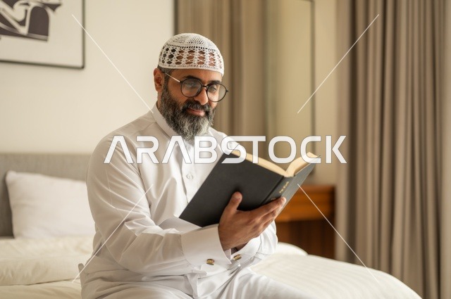 قضاء أوقات الفراغ بالتثقيف والتعلم, تنمية مهارات القراءة, الاهتمام بتصفح الكتب المفيدة، رجل عربي خليجي سعودي يرتدي الثوب التقليدي والقبعة البيضاء يجلس على السرير في المنزل ويحمل بيده كتاب