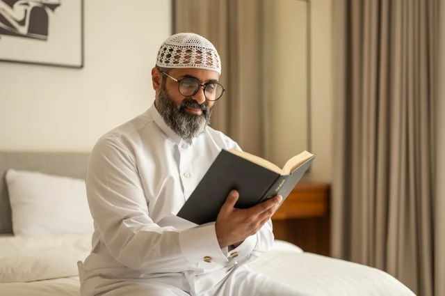 قضاء أوقات الفراغ بالتثقيف والتعلم, تنمية مهارات القراءة, الاهتمام بتصفح الكتب المفيدة، رجل عربي خليجي سعودي يرتدي الثوب التقليدي والقبعة البيضاء يجلس على السرير في المنزل ويحمل بيده كتاب
