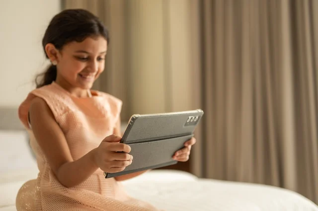 The concept of e-learning, following lessons remotely via the Internet, educational scientific curricula, a Saudi Arabian Gulf girl wearing a dress sitting on the bed holding a tablet in her hand, using a modern and advanced technical device