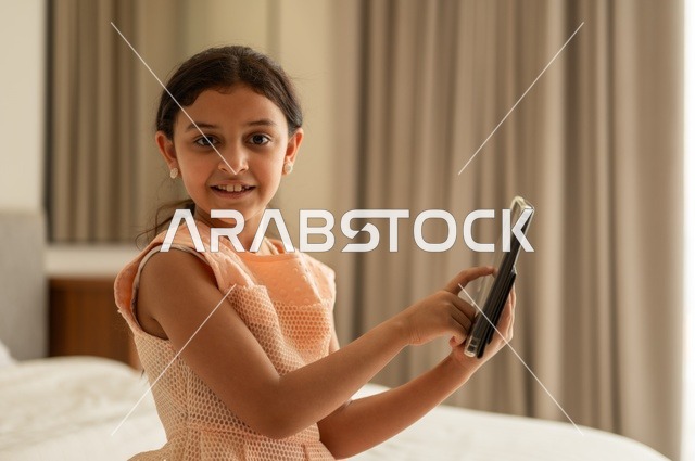 مفهوم التعليم الالكتروني، متابعة الدروس عن بعد عن طريق الانترنت، المناهج العلمية التعليمية،  فتاة عربية خليجية سعودية ترتدي الفستان تجلس على السرير تحمل بيدها الجهاز اللوحي، استخدام جهاز تقني حديث ومتطور
