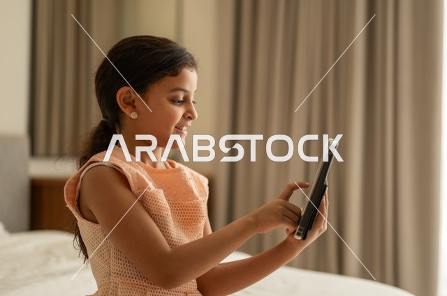 The concept of e-learning, following lessons remotely via the Internet, educational scientific curricula, a Saudi Arabian Gulf girl wearing a dress sitting on the bed holding a tablet in her hand, using a modern and advanced technical device
