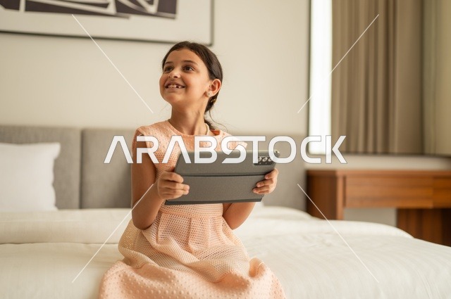 The concept of e-learning, following lessons remotely via the Internet, educational scientific curricula, a Saudi Arabian Gulf girl wearing a dress sitting on the bed holding a tablet in her hand, using a modern and advanced technical device