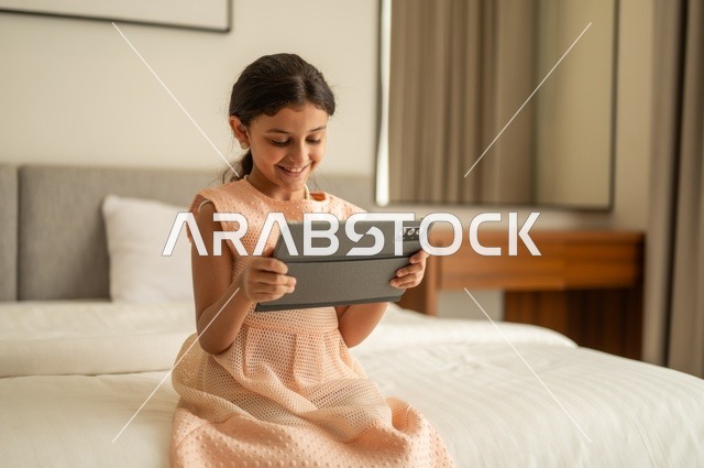 The concept of e-learning, following lessons remotely via the Internet, educational scientific curricula, a Saudi Arabian Gulf girl wearing a dress sitting on the bed holding a tablet in her hand, using a modern and advanced technical device