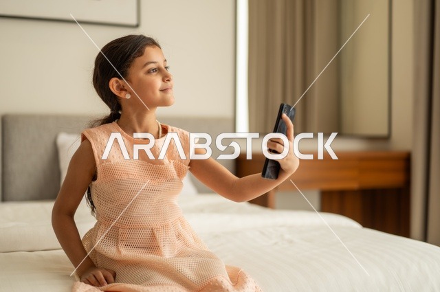 Chatting and communicating with family and friends, following lessons remotely via the Internet, browsing social networking sites, a Saudi Arabian Gulf girl wearing a dress sitting on the bed holding a mobile phone in her hand, using a modern and advanced technical device