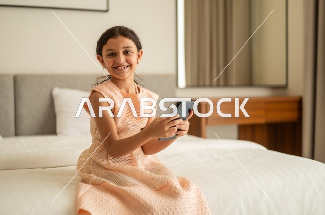 الدردشة والتواصل مع الاهل والأصدقاء، متابعة الدروس عن بعد عن طريق الانترنت، تصفح مواقع التواصل الاجتماعي،  فتاة عربية خليجية سعودية ترتدي الفستان تجلس على السرير تحمل بيدها الهاتف المحمول، استخدام جهاز تقني حديث ومتطور
