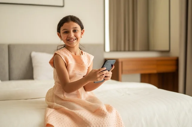 الدردشة والتواصل مع الاهل والأصدقاء، متابعة الدروس عن بعد عن طريق الانترنت، تصفح مواقع التواصل الاجتماعي،  فتاة عربية خليجية سعودية ترتدي الفستان تجلس على السرير تحمل بيدها الهاتف المحمول، استخدام جهاز تقني حديث ومتطور