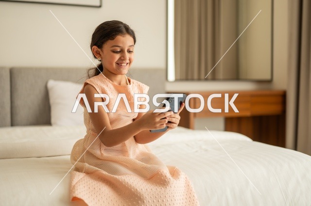 الدردشة والتواصل مع الاهل والأصدقاء، متابعة الدروس عن بعد عن طريق الانترنت، تصفح مواقع التواصل الاجتماعي،  فتاة عربية خليجية سعودية ترتدي الفستان تجلس على السرير تحمل بيدها الهاتف المحمول، استخدام جهاز تقني حديث ومتطور