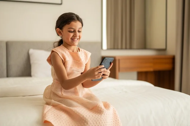 الدردشة والتواصل مع الاهل والأصدقاء، متابعة الدروس عن بعد عن طريق الانترنت، تصفح مواقع التواصل الاجتماعي،  فتاة عربية خليجية سعودية ترتدي الفستان تجلس على السرير تحمل بيدها الهاتف المحمول، استخدام جهاز تقني حديث ومتطور