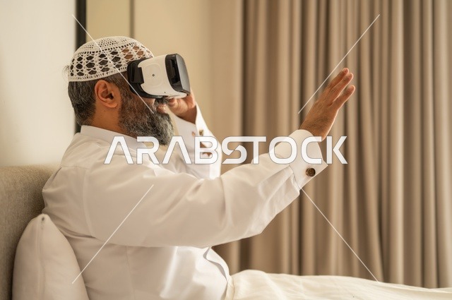 التفاعل والاندماج ومحاكاة العالم الخيالي، رجل عربي خليجي سعودي مبتسم يرتدي الثوب التقليدي والقبعة البيضاء بجلس على السرير يضع نظارة الواقع الافتراضي، المتعة والتسلية بالمشاهدة عبر نظارات ثلاثية الابعاد