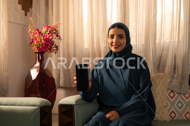 عرض شاشة سوداء فارغة عبر الجوال، الدردشه مع الأهل والأصدقاء، تصفح تطبيقات وبرامج التواصل الاجتماعي، إيماءات السرور والسعادة، سيدة عربية خليجية سعودية محجبة مبتسمة ترتدي عباءة تجلس على اريكة تمسك بيدها الهاتف المحمول، استخدام الاجهزة التقنية الحديثة والمتطورة