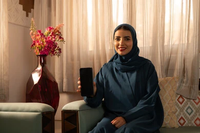 عرض شاشة سوداء فارغة عبر الجوال، الدردشه مع الأهل والأصدقاء، تصفح تطبيقات وبرامج التواصل الاجتماعي، إيماءات السرور والسعادة، سيدة عربية خليجية سعودية محجبة مبتسمة ترتدي عباءة تجلس على اريكة تمسك بيدها الهاتف المحمول، استخدام الاجهزة التقنية الحديثة والمتطورة