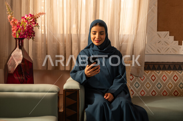 الدردشه مع الأهل والأصدقاء، تصفح تطبيقات وبرامج التواصل الاجتماعي، إيماءات السرور والسعادة، سيدة عربية خليجية سعودية محجبة مبتسمة ترتدي عباءة تجلس على اريكة تمسك بيدها الهاتف المحمول، استخدام الاجهزة التقنية الحديثة والمتطورة