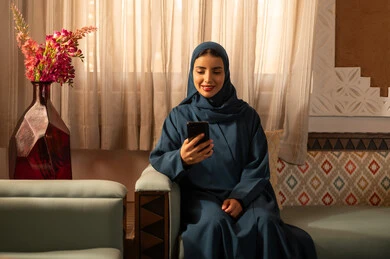 الدردشه مع الأهل والأصدقاء، تصفح تطبيقات وبرامج التواصل الاجتماعي، إيماءات السرور والسعادة، سيدة عربية خليجية سعودية محجبة مبتسمة ترتدي عباءة تجلس على اريكة تمسك بيدها الهاتف المحمول، استخدام الاجهزة التقنية الحديثة والمتطورة