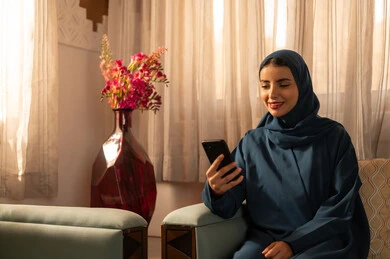 الدردشه مع الأهل والأصدقاء، تصفح تطبيقات وبرامج التواصل الاجتماعي، إيماءات السرور والسعادة، سيدة عربية خليجية سعودية محجبة مبتسمة ترتدي عباءة تجلس على اريكة تمسك بيدها الهاتف المحمول، استخدام الاجهزة التقنية الحديثة والمتطورة