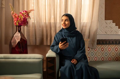 Chatting with family and friends, browsing social media applications and programs, gestures of joy and happiness, a smiling Saudi Arabian Gulf woman wearing a hijab and an abaya sitting on a sofa holding a mobile phone in her hand, using modern and advanced technological devices.