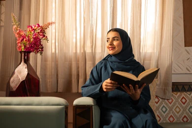 الاستمتاع بهواية مطالعة الكتب والقصص والروايات، ثقافة التعلم الذاتي، قضاء الوقت بالقراءه والتثقف، امرأة عربية خليجية سعودية محجبة ترتدي العباءة تجلس على اريكه مريحه تحمل بيديها كتاب