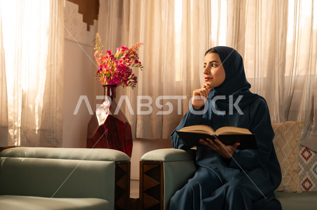 ثقافة التعلم الذاتي، قضاء الوقت بالقراءه والتثقف، الاستمتاع بهواية مطالعة الكتب والقصص والروايات، امرأة عربية خليجية سعودية محجبة ترتدي العباءة تجلس على اريكه مريحه تحمل بيديها كتاب