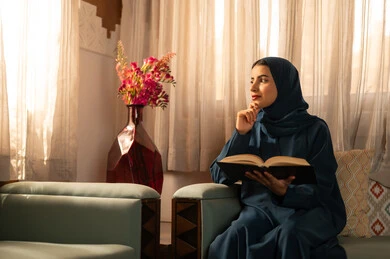 ثقافة التعلم الذاتي، قضاء الوقت بالقراءه والتثقف، الاستمتاع بهواية مطالعة الكتب والقصص والروايات، امرأة عربية خليجية سعودية محجبة ترتدي العباءة تجلس على اريكه مريحه تحمل بيديها كتاب