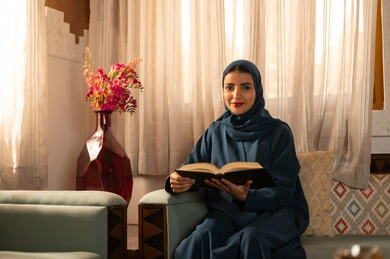 The culture of self-learning, spending time reading and educating oneself, enjoying the hobby of reading books, stories, and novels. A Saudi Arabian Gulf woman wearing a hijab and an abaya sits on a comfortable couch holding a book in her hands.