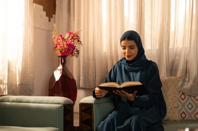 Enjoying the hobby of reading books, stories, and novels, the culture of self-learning, spending time reading and gaining knowledge, a Gulf Arab Saudi woman wearing a hijab and an abaya sits on a comfortable sofa holding a book in her hands.
