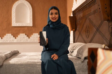 Chatting with family and friends, displaying a blank black screen on the mobile, browsing social media applications and programs, gestures of joy and happiness, a smiling Saudi Arabian Gulf woman wearing a hijab and an abaya sitting on the bed holding a mobile phone in her hand, using modern and advanced technological devices.
