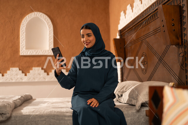 الدردشه مع الأهل والأصدقاء،  تصفح تطبيقات وبرامج التواصل الاجتماعي، إيماءات السرور والسعادة، سيدة عربية خليجية سعودية محجبة مبتسمة ترتدي عباءة تجلس على السرير تمسك بيدها الهاتف المحمول، استخدام الاجهزة التقنية الحديثة والمتطورة
