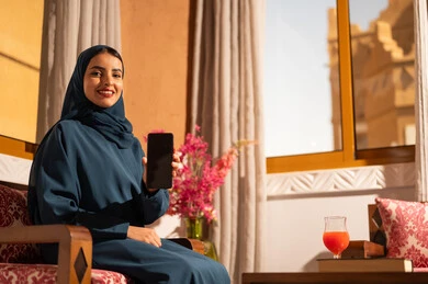 Displaying a blank black screen on the mobile, chatting with family and friends, browsing social media applications and programs, gestures of joy and happiness, a smiling Saudi Arabian Gulf woman wearing a hijab and an abaya sitting on a sofa holding a mobile phone in her hand, using modern and advanced technological devices.