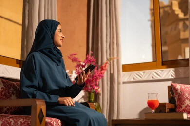 الدردشه مع الأهل والأصدقاء، تصفح تطبيقات وبرامج التواصل الاجتماعي، إيماءات السرور والسعادة، سيدة عربية خليجية سعودية محجبة مبتسمة ترتدي عباءة تجلس على اريكة تمسك بيدها الهاتف المحمول، استخدام الاجهزة التقنية الحديثة والمتطورة