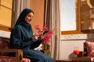 Chatting with family and friends, browsing social media applications and programs, gestures of joy and happiness, a smiling Saudi Arabian Gulf woman wearing a hijab and an abaya sitting on a sofa holding a mobile phone in her hand, using modern and advanced technological devices.