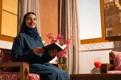 The culture of self-learning, spending time reading and educating oneself, enjoying the hobby of reading books, stories, and novels. A Saudi Arabian Gulf woman wearing a hijab and an abaya sits on a comfortable couch holding a book in her hands.
