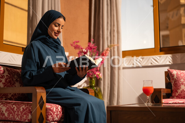 The culture of self-learning, spending time reading and educating oneself, enjoying the hobby of reading books, stories, and novels. A Saudi Arabian Gulf woman wearing a hijab and an abaya sits on a comfortable couch holding a book in her hands.