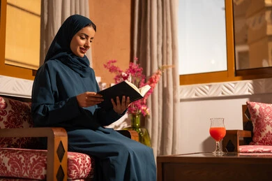 The culture of self-learning, spending time reading and educating oneself, enjoying the hobby of reading books, stories, and novels. A Saudi Arabian Gulf woman wearing a hijab and an abaya sits on a comfortable couch holding a book in her hands.