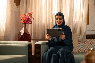 العمل عن بعد عبر الانترنت، الدردشه مع الأهل والأصدقاء،  تصفح تطبيقات وبرامج التواصل الاجتماعي، إيماءات السرور والسعادة، سيدة عربية خليجية سعودية محجبة مبتسمة ترتدي عباءة تجلس على الاريكة تمسك بيدها التابليت المحمول، استخدام الاجهزة التقنية الحديثة والمتطورة