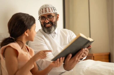 Spending free time in education and learning, developing reading skills, interest in browsing useful books, a Saudi Arabian Gulf man wearing traditional clothes and a white hat sitting on the bed with his daughter at home and holding a book in his hand