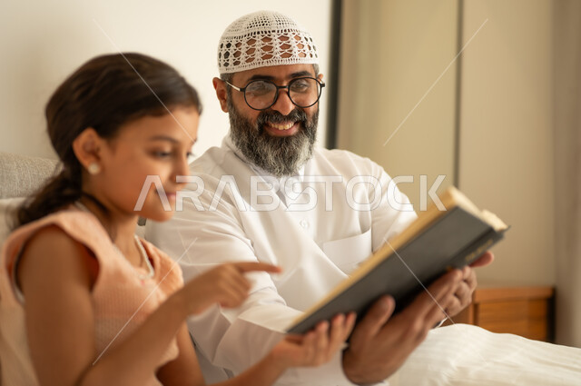 قضاء أوقات الفراغ بالتثقيف والتعلم, تنمية مهارات القراءة, الاهتمام بتصفح الكتب المفيدة، رجل عربي خليجي سعودي يرتدي الثوب التقليدي والقبعة البيضاء يجلس على السرير مع ابنته في المنزل ويحمل بيده كتاب
