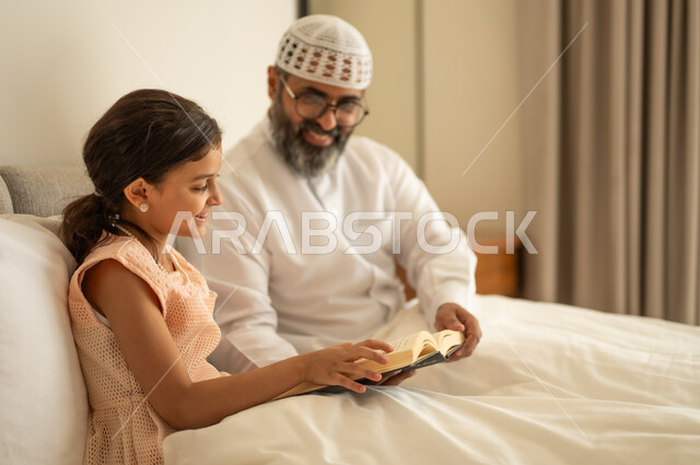 Spending free time in education and learning, developing reading skills, interest in browsing useful books, a Saudi Arabian Gulf man wearing traditional clothes and a white hat sitting on the bed with his daughter at home and holding a book in his hand