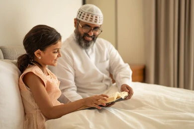 قضاء أوقات الفراغ بالتثقيف والتعلم, تنمية مهارات القراءة, الاهتمام بتصفح الكتب المفيدة، رجل عربي خليجي سعودي يرتدي الثوب التقليدي والقبعة البيضاء يجلس على السرير مع ابنته في المنزل ويحمل بيده كتاب