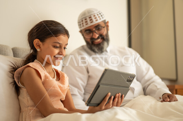 Spending free time in education and learning, developing reading skills, interest in browsing useful books, a Saudi Arabian Gulf man wearing traditional clothes and a white hat sitting on the bed with his daughter at home and holding a book in his hand