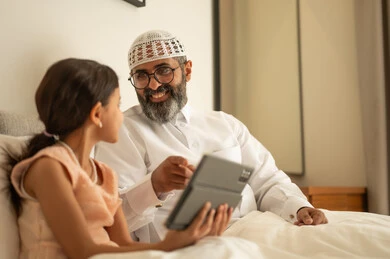 Spending free time in education and learning, developing reading skills, interest in browsing useful books, a Saudi Arabian Gulf man wearing traditional clothes and a white hat sitting on the bed with his daughter at home and holding a book in his hand