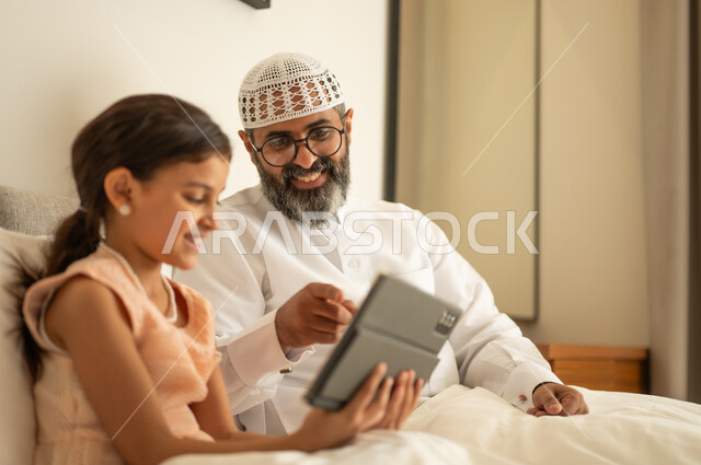 Spending free time in education and learning, developing reading skills, interest in browsing useful books, a Saudi Arabian Gulf man wearing traditional clothes and a white hat sitting on the bed with his daughter at home and holding a book in his hand