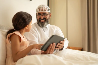 مفهوم التعليم الالكتروني، متابعة الدروس عن بعد عن طريق الانترنت، المناهج العلمية التعليمية،  رجل عربي خليجي سعودي يرتدي الثوب التقليدي والقبعة البيضاء يجلس على السرير مع ابنته، استخدام الجهاز اللوحي، استعمال جهاز تقني حديث ومتطور