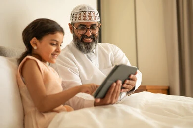 مفهوم التعليم الالكتروني، متابعة الدروس عن بعد عن طريق الانترنت، المناهج العلمية التعليمية،  رجل عربي خليجي سعودي يرتدي الثوب التقليدي والقبعة البيضاء يجلس على السرير مع ابنته، استخدام الجهاز اللوحي، استعمال جهاز تقني حديث ومتطور