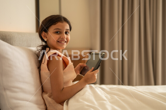 مفهوم التعليم الالكتروني، متابعة الدروس عن بعد عن طريق الانترنت، المناهج العلمية التعليمية،  فتاة عربية خليجية سعودية ترتدي الفستان تجلس على السرير تحمل بيدها الجهاز اللوحي، استخدام جهاز تقني حديث ومتطور