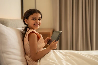 مفهوم التعليم الالكتروني، متابعة الدروس عن بعد عن طريق الانترنت، المناهج العلمية التعليمية،  فتاة عربية خليجية سعودية ترتدي الفستان تجلس على السرير تحمل بيدها الجهاز اللوحي، استخدام جهاز تقني حديث ومتطور