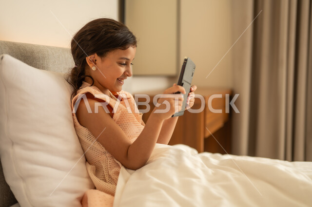 The concept of e-learning, following lessons remotely via the Internet, educational scientific curricula, a Saudi Arabian Gulf girl wearing a dress sitting on the bed holding a tablet in her hand, using a modern and advanced technical device