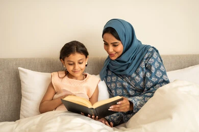 قراءة القصص قبل النوم، ثقافة التعلم الذاتي، قضاء الوقت بالقراءه والتثقف، الاستمتاع بهواية مطالعة الكتب والروايات، امرأة عربية خليجية سعودية محجبة ترتدي العباءة تجلس على السرير مع ابنتها تحمل بيديها كتاب