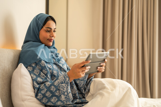 Working remotely via the Internet, chatting with family and friends, browsing social media applications and programs, a smiling veiled Saudi Arabian Gulf woman wearing an abaya sitting on the bed holding a tablet in her hand, using modern and advanced technical devices