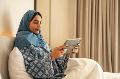 العمل عن بعد عبر الانترنت، الدردشه مع الأهل والأصدقاء،  تصفح تطبيقات وبرامج التواصل الاجتماعي، سيدة عربية خليجية سعودية محجبة مبتسمة ترتدي عباءة تجلس على السرير تمسك بيدها التابليت المحمول، استخدام الاجهزة التقنية الحديثة والمتطورة