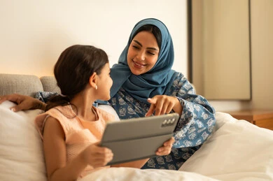 المناهج العلمية التعليمية،  فتاة عربية خليجية سعودية ترتدي الفستان تحمل بيدها الجهاز اللوحي، استخدام جهاز تقني حديث ومتطور، ام ترتدي العباءة تجلس مع ابتنها على السرير، مفهوم التعليم الالكتروني، متابعة الدروس عن بعد عن طريق الانترنت